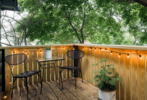 Cozy balcony with two chairs, a small table, string lights, and a potted plant surrounded by greenery.