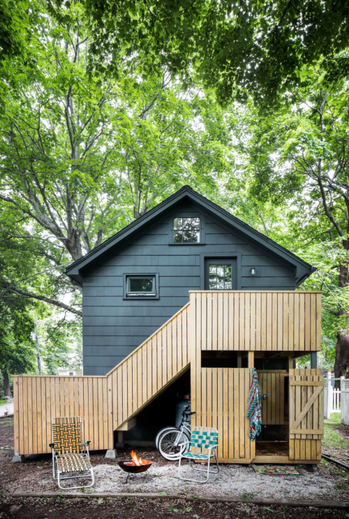 A modern two-story house with a wooden deck, surrounded by trees, featuring a fire pit and outdoor seating.