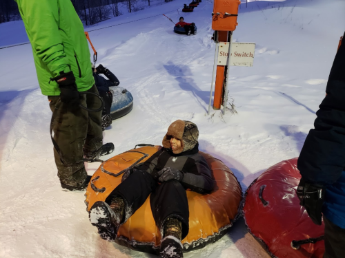A child sits in a snow tube, ready to go down a snowy hill, while adults prepare nearby. Snowy landscape in the background.