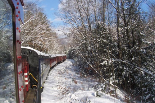 A snowy landscape with a train winding through trees, viewed from the side of a train car.