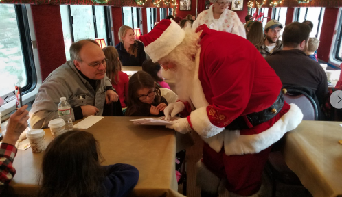 Santa Claus interacts with a young girl at a festive train event, surrounded by families enjoying the holiday atmosphere.