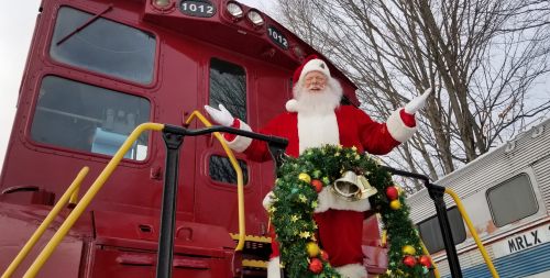 Santa Claus stands on a red train engine, smiling and welcoming with a festive wreath around a bell.