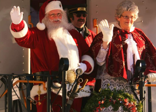 Santa Claus and Mrs. Claus wave cheerfully from a festive train, surrounded by holiday decorations.