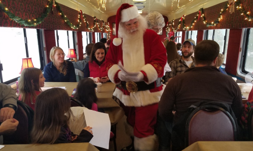 A festive train interior with Santa interacting with children and families, decorated with holiday lights and garlands.