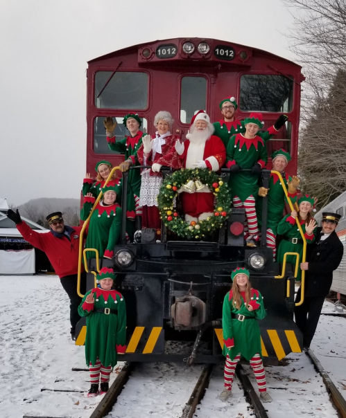 A festive group of elves and Santa pose on a train, surrounded by snow and holiday decorations.