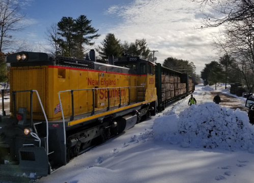 A yellow train with wooden cargo cars moves through a snowy landscape, surrounded by trees and a clear blue sky.