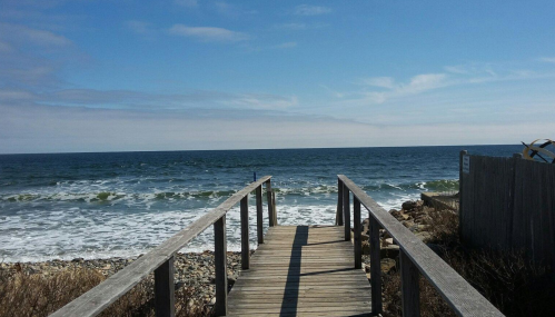 A wooden walkway leads to a calm ocean with gentle waves under a clear blue sky.