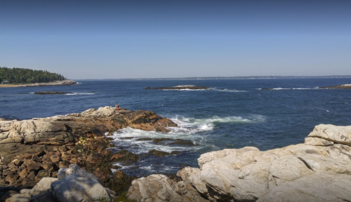 A rocky coastline with gentle waves, under a clear blue sky and distant trees along the shore.