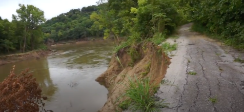 A winding, cracked road alongside a river, with eroded banks and lush greenery on either side.