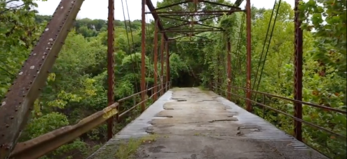 An old, rusty bridge surrounded by lush green trees and overgrown vegetation, leading into a dense forest.