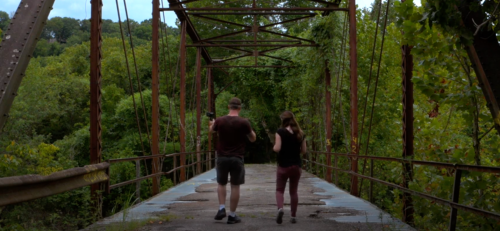 Two people walk hand in hand on an old, rusty bridge surrounded by lush greenery.