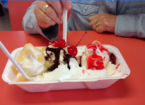 A person holds a dish of ice cream topped with cherries, chocolate sauce, and whipped cream on a red table.