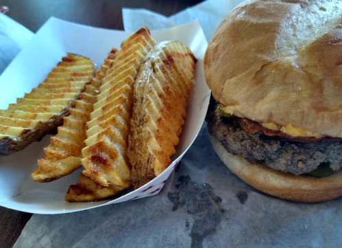 A close-up of a burger with cheese and pickles, accompanied by a side of crispy waffle fries on a paper tray.