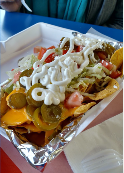 A tray of nachos topped with cheese, jalapeños, lettuce, tomatoes, and sour cream, served in a foil container.