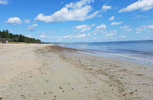A serene beach scene with soft sand, calm water, and scattered clouds in a blue sky.