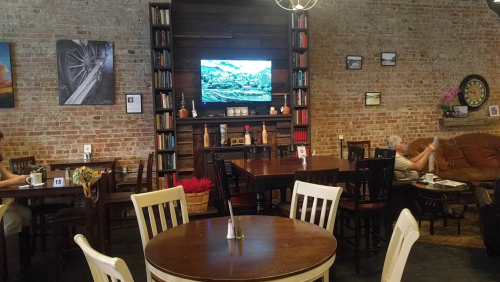 Cozy café interior with wooden tables, a TV on the wall, and a bookshelf, featuring a few patrons enjoying their time.