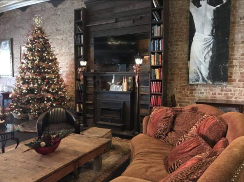 Cozy living room with a Christmas tree, fireplace, bookshelves, and a large brown sofa. Warm, festive atmosphere.