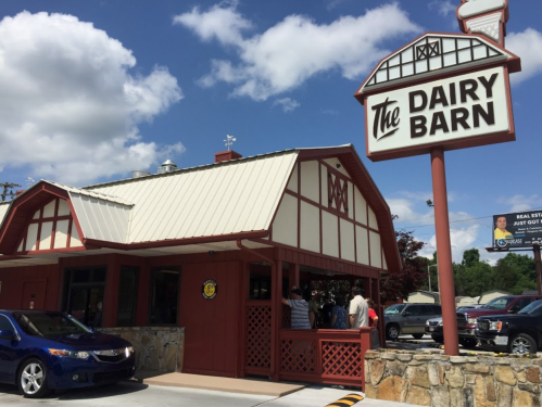 A red and white building with a sign reading "The Dairy Barn," featuring a car parked in front and a blue sky above.