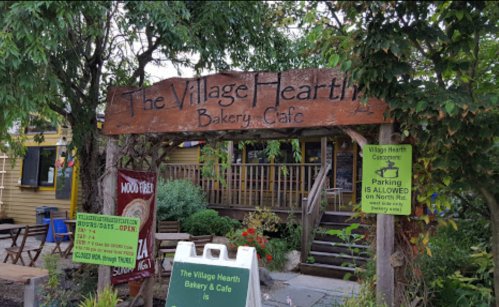 Sign for "The Village Hearth Bakery Cafe" with outdoor seating and greenery surrounding the entrance.