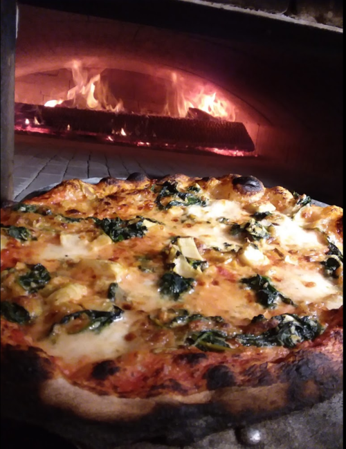 A freshly baked pizza with spinach and cheese, placed in a wood-fired oven with flames in the background.