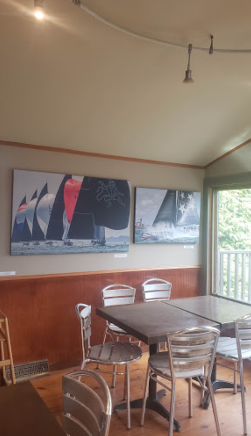 A cozy interior with wooden walls, featuring three large sailing-themed artworks and metal chairs around tables.