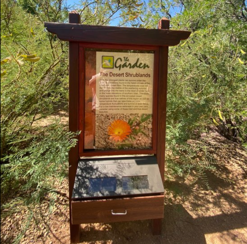 Sign for "The Desert Shrublands" exhibit in a garden, surrounded by greenery and desert plants.