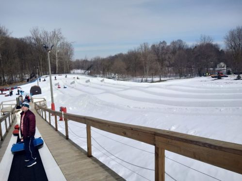 A snowy tubing hill with people enjoying the slopes, viewed from a wooden walkway. Trees line the background.