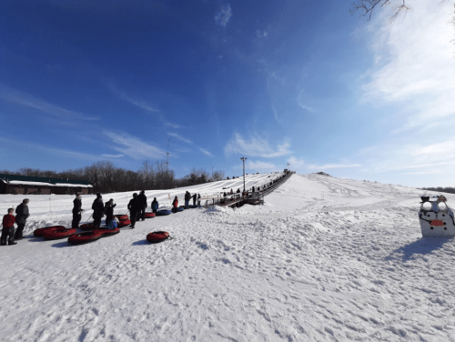 A snowy hill with people waiting in line to go tubing, under a clear blue sky.
