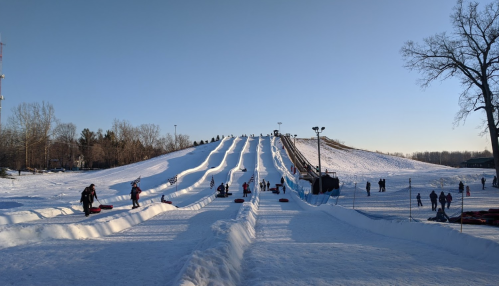 A snowy hill with multiple tubing lanes, people sliding down and walking up, under a clear blue sky.