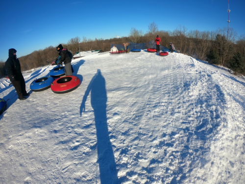 People preparing to go tubing on a snowy hill, with a clear blue sky in the background. Shadows cast on the snow.