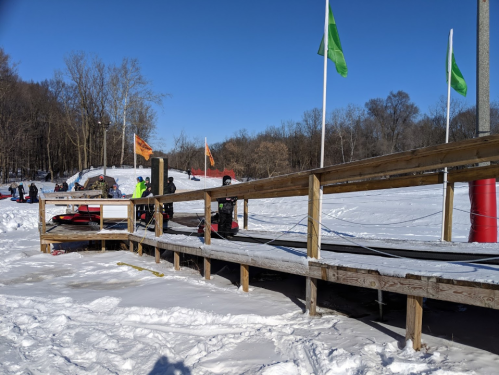 A snowy landscape with a wooden dock, flags flying, and people preparing snowmobiles in the background.