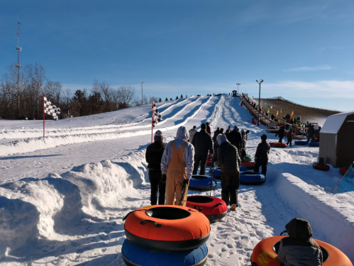 A line of people waiting to go tubing down a snowy hill on a clear day, with colorful tubes lined up nearby.