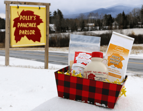 A basket with pancake mix, maple syrup, and sugar in front of Polly's Pancake Parlor sign and snowy landscape.