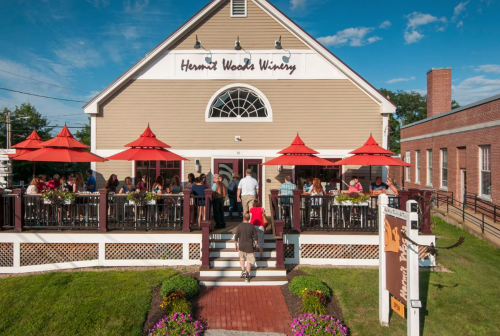 A winery with outdoor seating, red umbrellas, and people enjoying drinks on a sunny day.