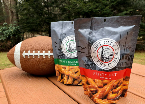 Two bags of Port City Pretzels, "Tasty Ranch" and "Feisty Hot," next to a football on a wooden surface outdoors.
