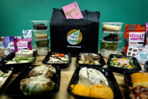 A variety of prepared meals and snacks displayed with a black bag labeled "all real meal" and a cheerful note saying "Yay!"