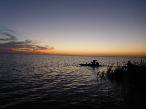 A serene sunset over a calm lake, with silhouettes of people kayaking near the shore.