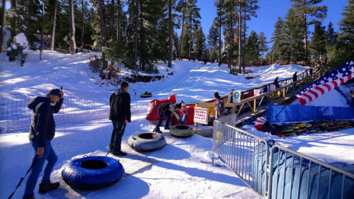 People preparing to go tubing on a snowy hill, with trees and a colorful ramp in the background.