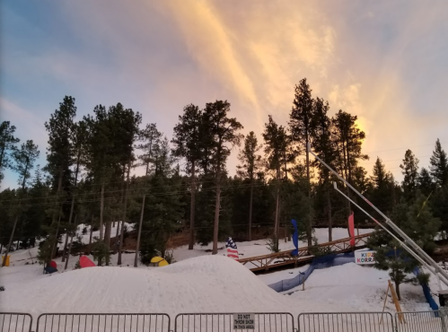 A snowy landscape with trees and a ski ramp under a colorful sunset sky. Flags are visible in the background.