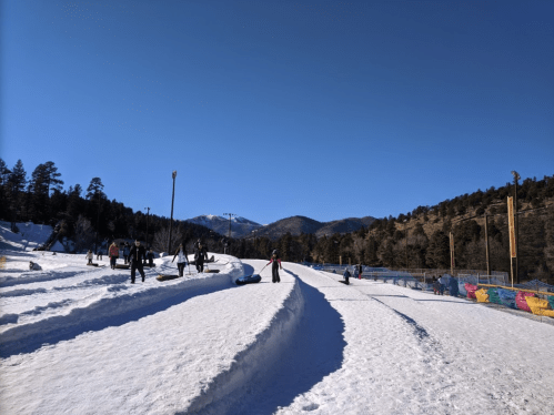 A snowy landscape with people enjoying winter activities under a clear blue sky and mountains in the background.