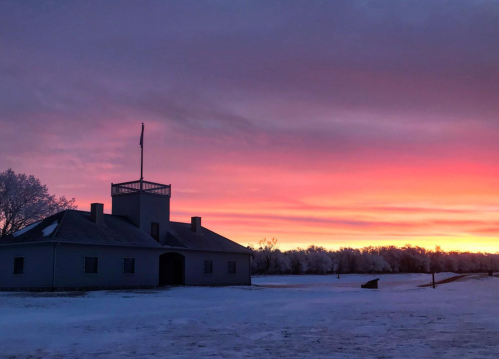 A historic building silhouetted against a vibrant pink and purple sunset, with snow covering the ground.