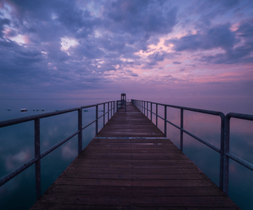 A wooden pier extends into calm waters under a colorful, cloudy sky at dawn.