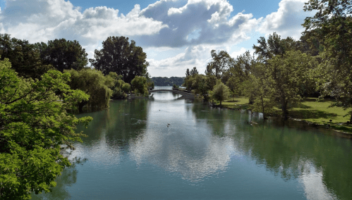 A serene river scene with lush greenery, a bridge in the distance, and fluffy clouds reflecting in the water.
