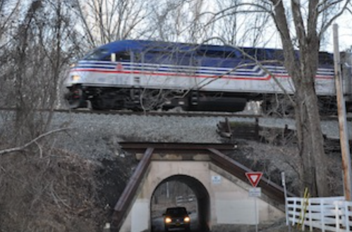 A train passes over a road under a bridge, surrounded by bare trees in a rural setting.