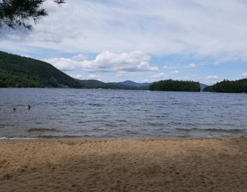 A sandy beach by a lake surrounded by green mountains and a cloudy blue sky, with a few people swimming.