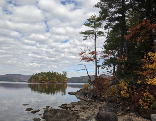 A serene lake scene with an island, surrounded by colorful autumn foliage and a cloudy sky.