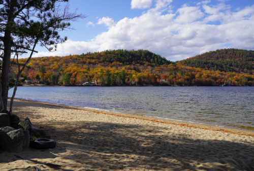 A serene lakeside view with sandy shore, colorful autumn trees on hills, and a partly cloudy sky.