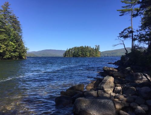 A serene lake scene with rocky shorelines, lush trees, and a clear blue sky in the background.
