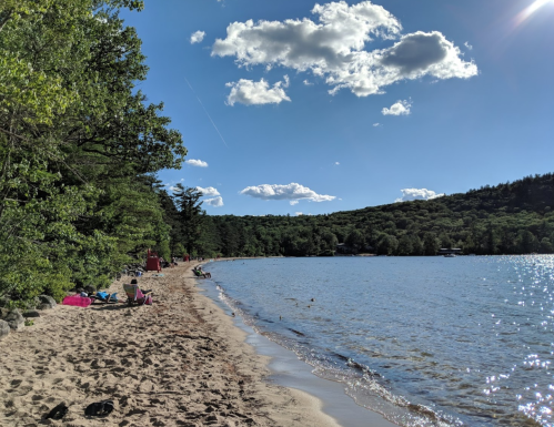A sandy beach by a calm lake, with trees and clouds in the background, and people relaxing along the shore.