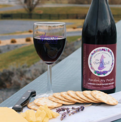 A glass of red wine beside a bottle labeled "Pardon My Purple," with cheese and crackers on a table outdoors.
